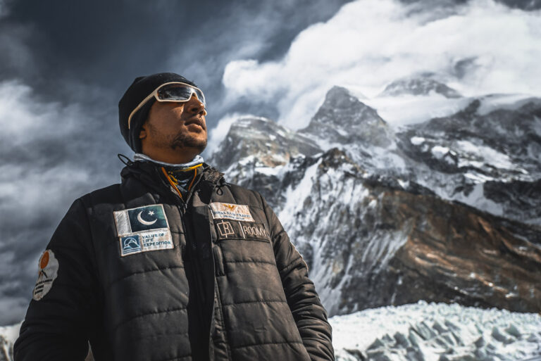 Sajid Sadpara in a black down suit with a Pakistan flag patch, K2 and a glacier behind him, winter 2021